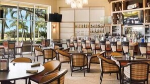 A spacious indoor dining area with multiple tables and chairs, large windows with a view of palm trees, and a bar area with shelves of bottles. - Greenlinks Luxury Condo, 3BD, ground floor, golf, pool (Naples)