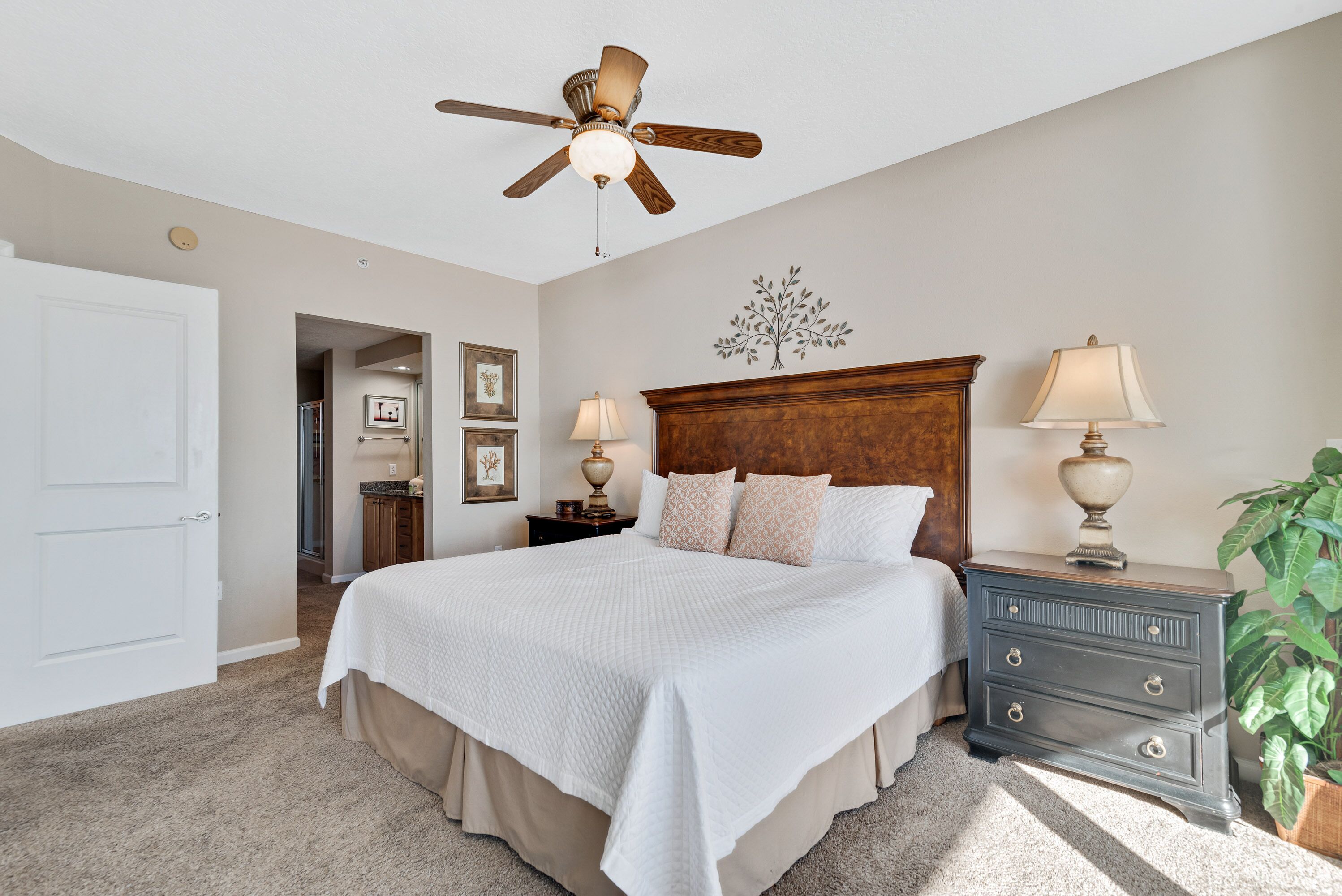A bedroom with a large bed, a wooden headboard, two bedside tables with lamps, a ceiling fan, and a door leading to another room.