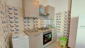 A compact kitchen with a gas stove, sink, and oven, light wooden cabinets, and floral patterned tiles. - Cortina 2-9 Two room seafront apartment with air conditioning (Comacchio)