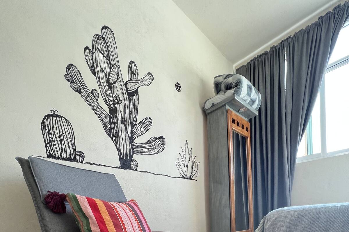 A room with a wooden-framed rocking chair, a bed with a grey blanket, a colorful striped rug, and a wall mural of cacti.