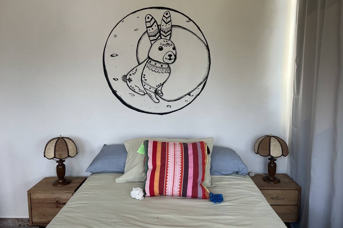 A bedroom with a bed, two nightstands, two lamps, a colorful striped pillow, and a wall decoration of a stylized animal.