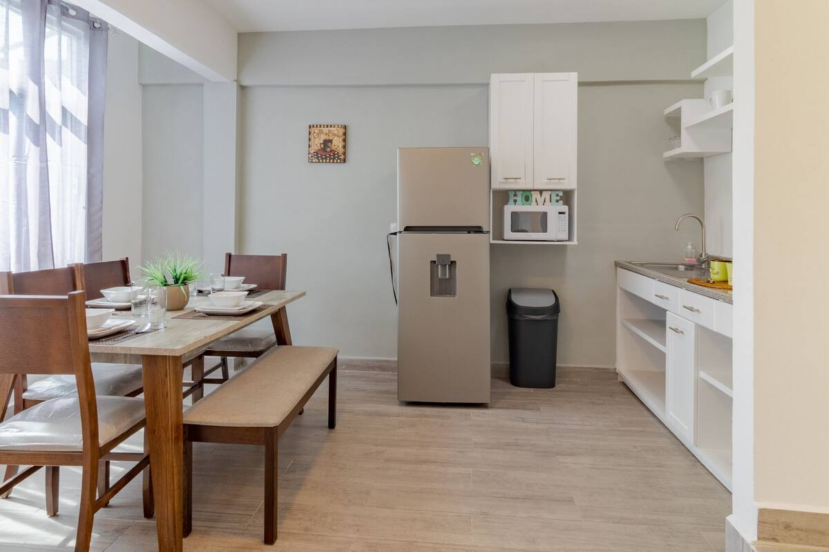A compact kitchen with a refrigerator, microwave, and white cabinets. There is a dining table with chairs and a bench.
