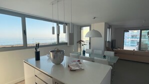 A modern kitchen with a view of the sea, a dining area with a glass table, and a living room with a sofa.