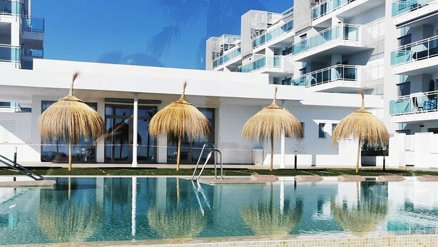 A swimming pool with a tiled edge, surrounded by a white building with balconies and thatched umbrellas.