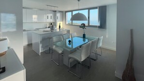 A modern kitchen with white cabinets, a dining area with a glass table and white chairs, and a large window with a view of the ocean.