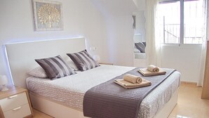 A neatly made bed with pillows, a bedside table, a mirror, and a window with a view. - Apartment with Large Roof Solarium, Pool, Garden, Barbecue, A/C & Wi-Fi (Orihuela Costa)