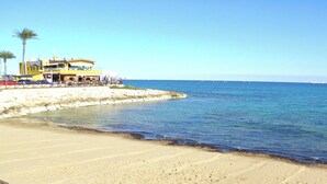 A beach with a building, palm trees, and a clear blue sea. - Apartment with Large Roof Solarium, Pool, Garden, Barbecue, A/C & Wi-Fi (Orihuela Costa)