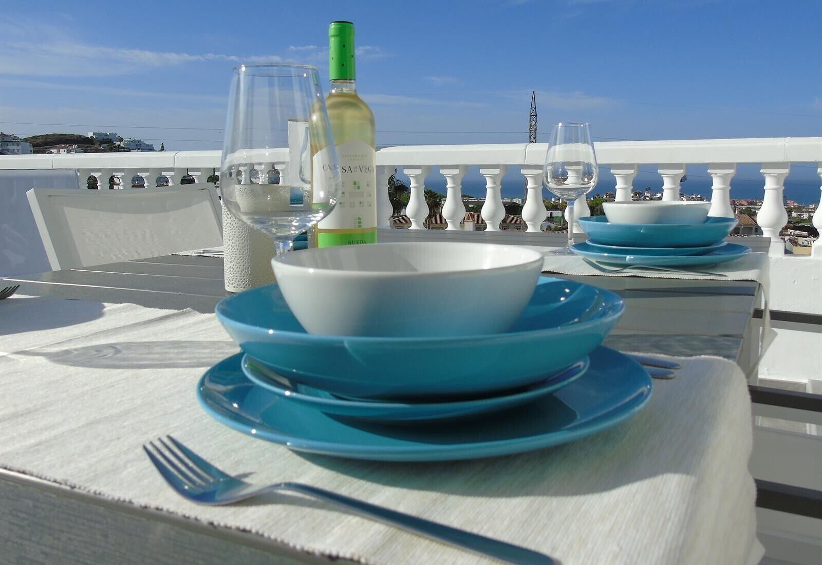 A table set with blue plates, a wine bottle, and glasses, overlooking a coastal view.