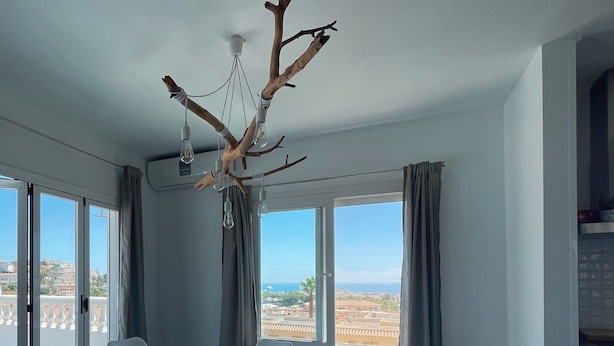 A dining area with a round table, white chairs, a branch chandelier, large windows, and a view of the landscape. - Apartment "Atico Montesol" with Sea View, shared Pool & Terrace (Torrox Costa)