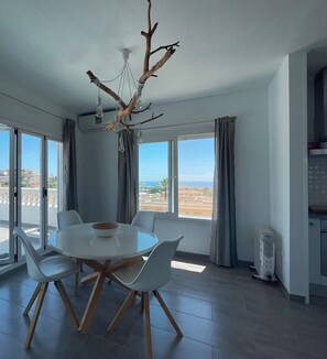 A dining area with a round table, white chairs, a branch chandelier, large windows, and a view of the landscape. - Apartment "Atico Montesol" with Sea View, shared Pool & Terrace (Torrox Costa)