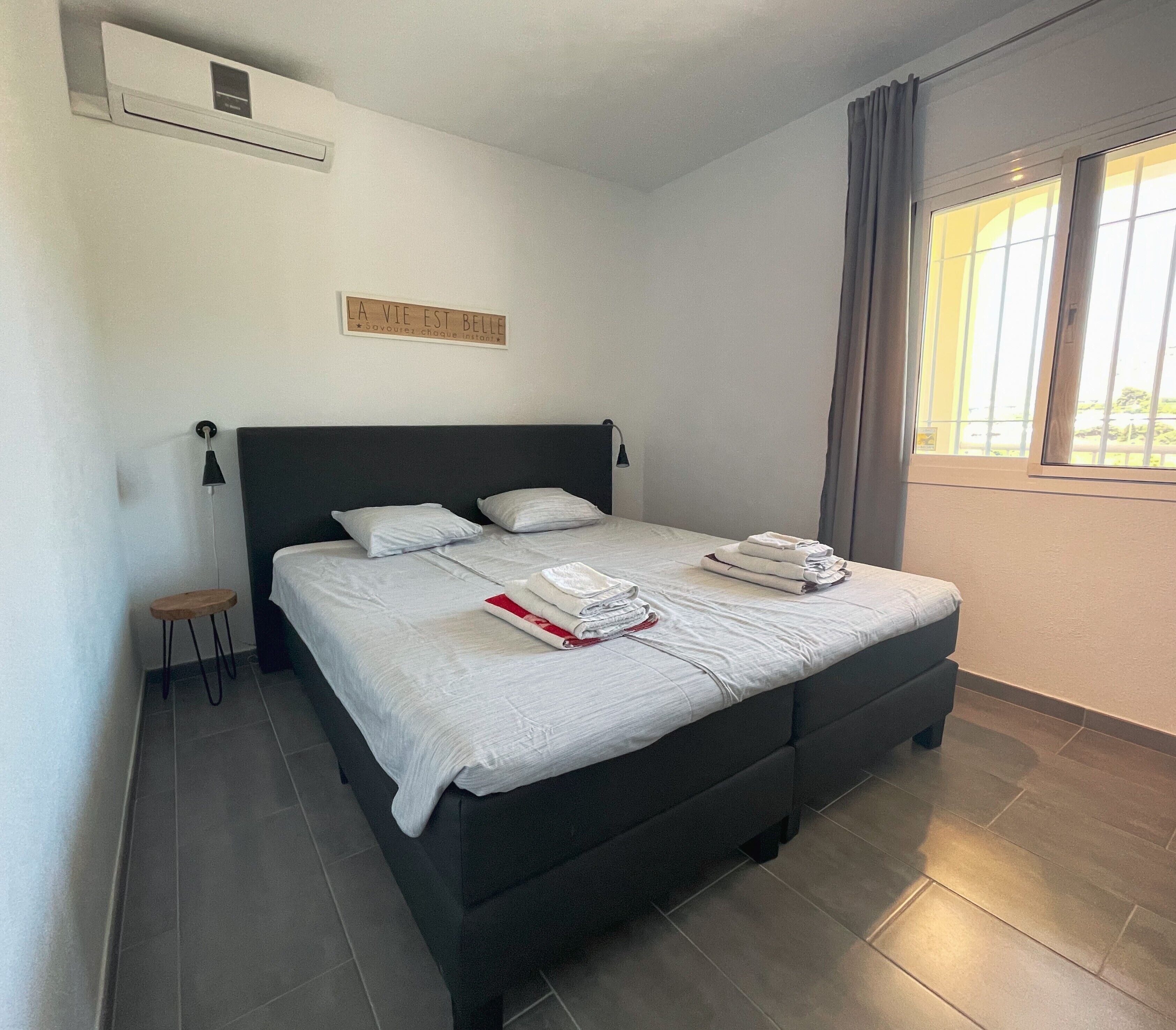 Un lit bien fait, avec des oreillers et des serviettes pliées, un petit tabouret et un climatiseur fixé au mur.