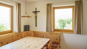 Een eetruimte met een houten tafel, bankjes, een kruis en uitzicht door grote ramen.