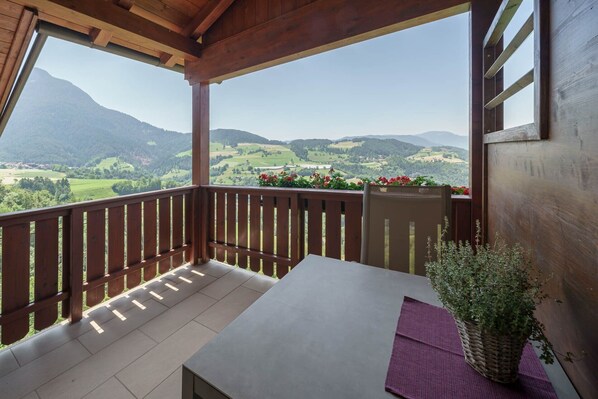 A balcony with a table and chairs, wooden railings, and a view of green hills and mountains.