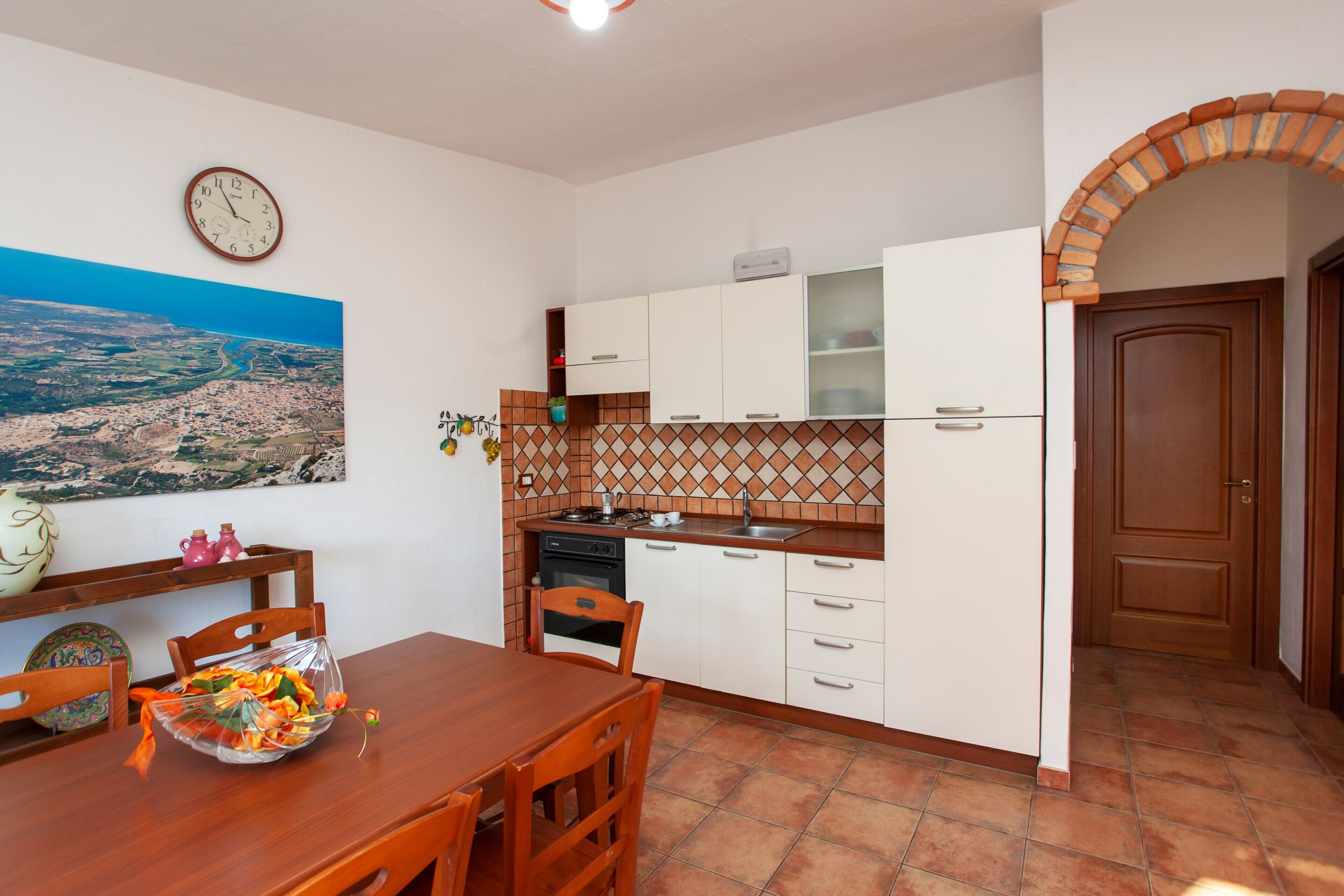 Una cocina con armarios blancos, salpicadero de azulejos, una mesa de comedor de madera y un reloj en la pared.