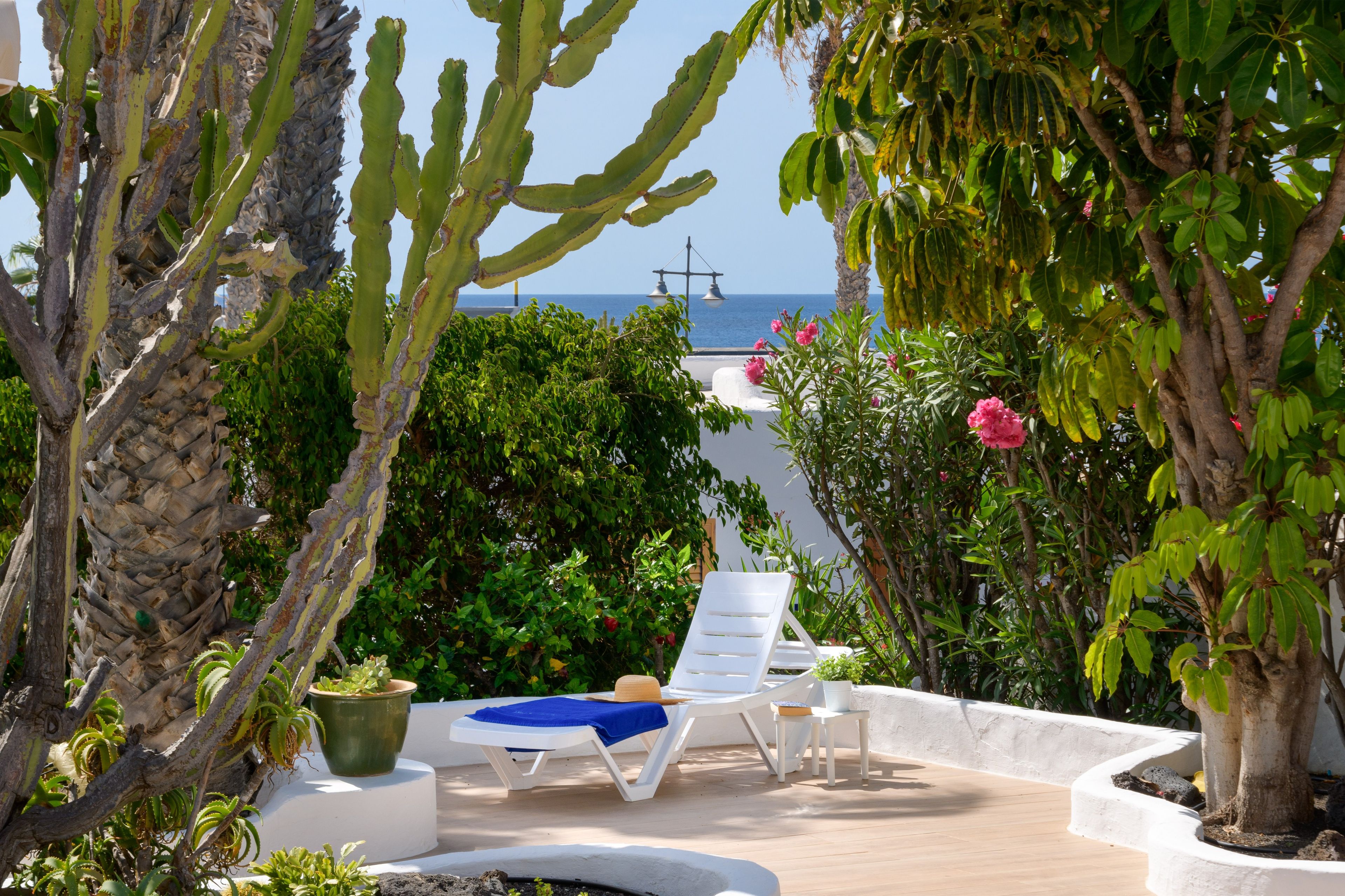 A sunlit outdoor seating area with a view of the sea, surrounded by various plants and a white fence.