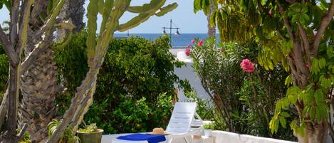 A sunlit outdoor seating area with a view of the sea, surrounded by various plants and a white fence.
