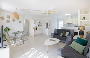 Uma sala de estar moderna com um sofá cinzento, uma mesa de centro e uma televisão. Há uma área de jantar com uma mesa e cadeiras de vidro. O quarto tem paredes brancas e um ventilador de teto.