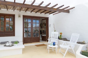 Une façade blanche avec des portes et des fenêtres en bois, un patio couvert avec deux chaises blanches et une petite table sur laquelle se trouvent une théière et une plante.