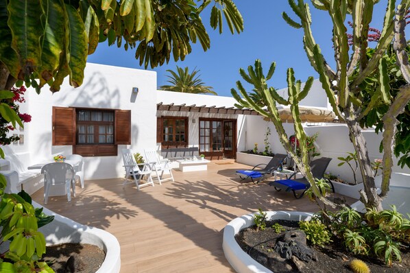 A sunlit patio with white walls, wooden shutters, and a variety of seating including chairs and a sofa.