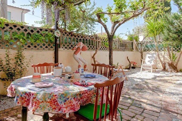 An outdoor dining area with a table set for two, surrounded by chairs, a wooden fence, and greenery.