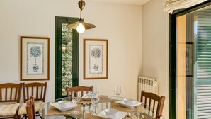 A dining area with a glass table, wooden chairs, framed artwork, and a window with blinds. - Family villa in Torremirona Resort (Navata)