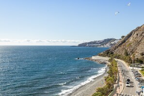 A coastal road with a beach, a rocky hillside, and a clear sky.