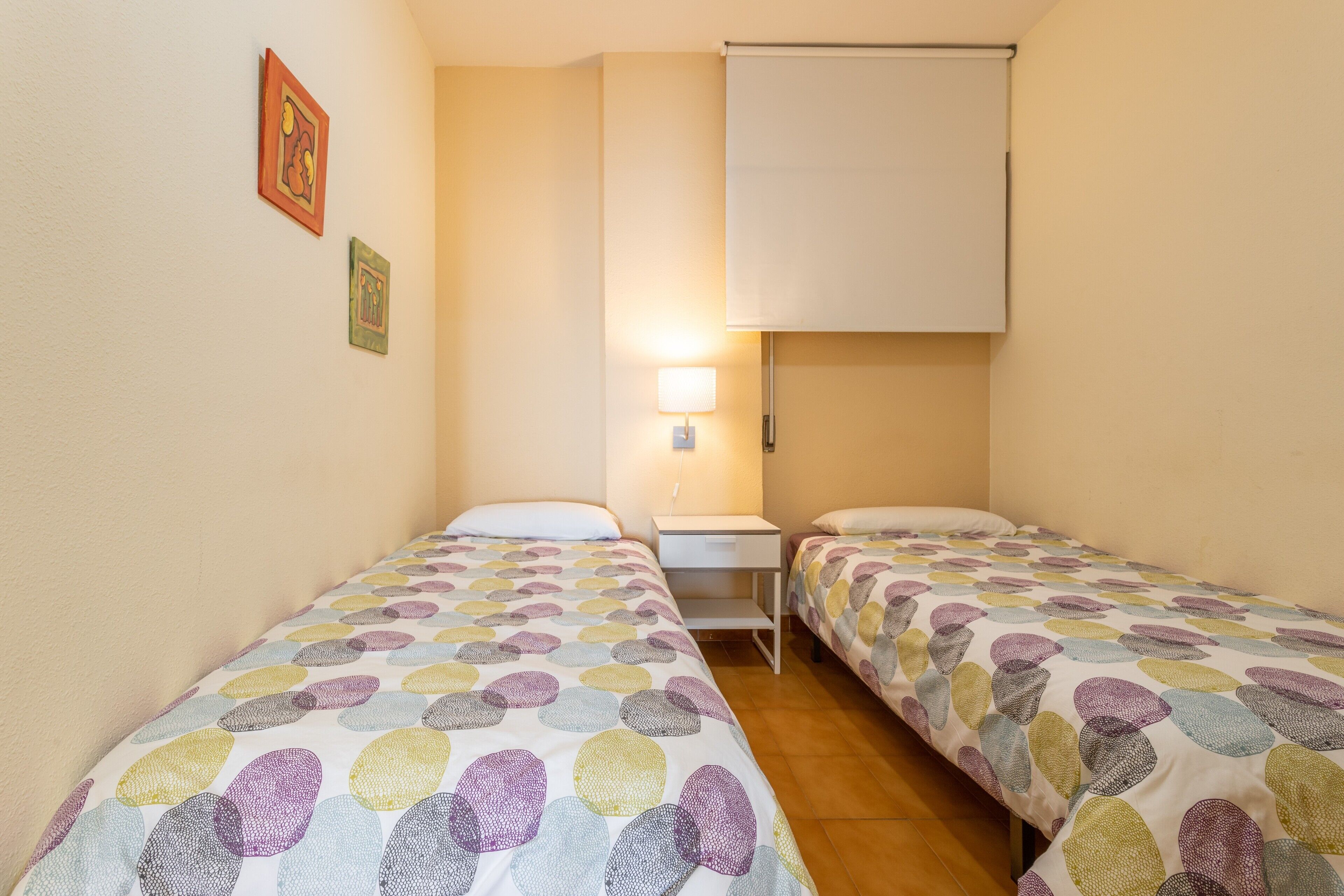Una habitación pequeña y limpia con dos camas individuales, una mesita de noche, una lámpara y un ventilador de techo blanco.