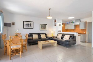 A living room with a sofa, coffee table, and dining set. There is a kitchen area with cabinets and a sink.