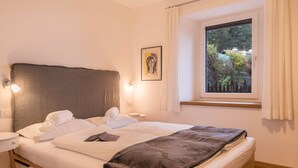 Un lit bien fait, avec des oreillers et une couverture, une table de chevet avec une lampe, un tableau encadré au mur et une fenêtre donnant sur des arbres et des immeubles.