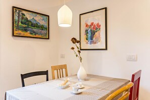 Ein Esstisch mit weißem Tischtuch, zwei Stühle, eine Vase mit Trockenblumen und zwei gerahmte Gemälde an der Wand.
