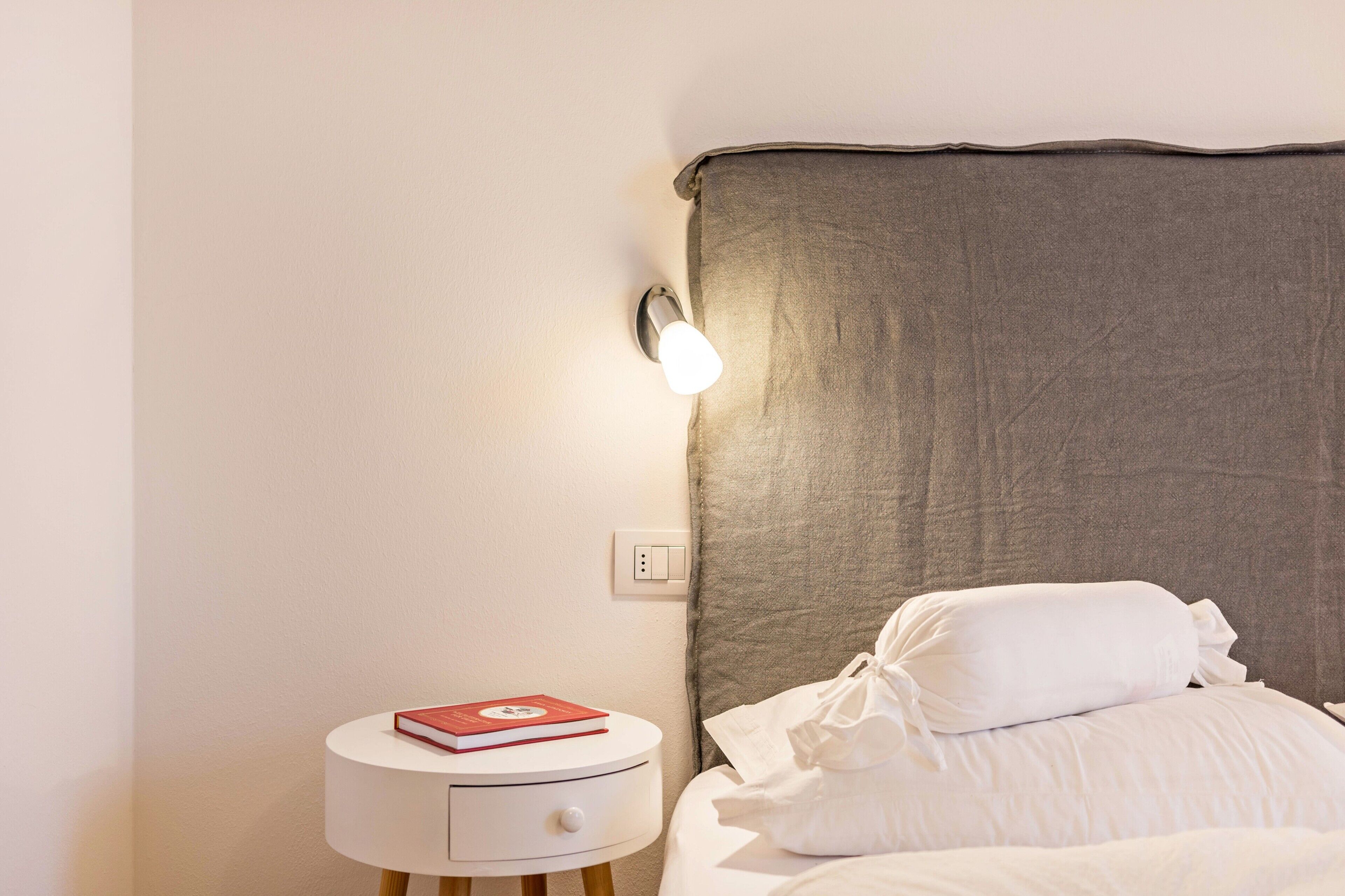A neatly made bed with white bedding, a bedside table with a book, and a wall-mounted light fixture.