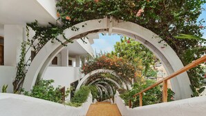 Una escalera con paredes blancas y barandilla de madera, que conduce a un espacio al aire libre con vegetación y vistas a edificios.
