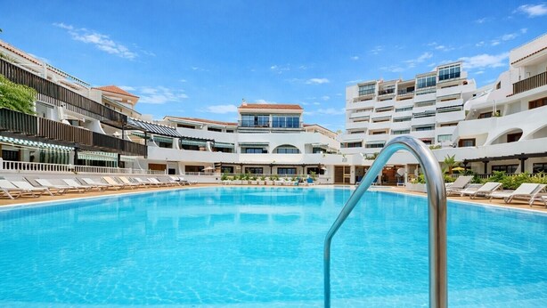 Una piscina con sillas de descanso, una escalera metálica para la piscina y edificios de varios pisos con balcones al fondo.