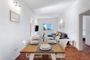 A dining area with a wooden table set for four, white chairs, a flat-screen TV mounted on the wall, and a sofa in the background. - Rural Country House "Finca Los Corcos" with 2 Pools, Garden, Mountain & Sea View (guia de Isora)