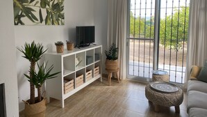 A living room with a TV on a white cabinet, a large window with a view of greenery, and a patterned ottoman.