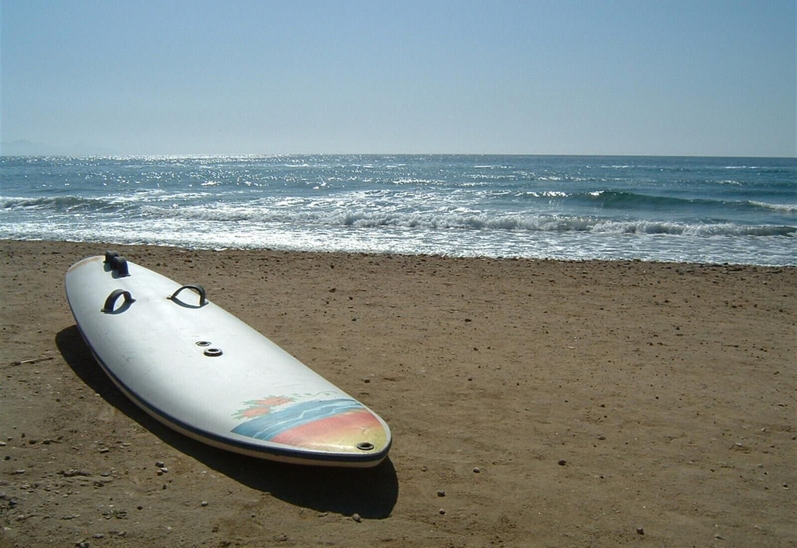 Una tabla de surf sobre arena con el océano al fondo.