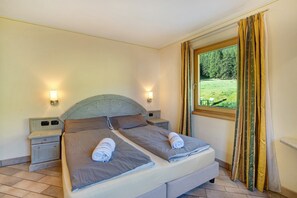 A double bed with two pillows and a blanket, a nightstand, a lamp, and a window with curtains.