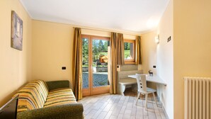 Une pièce avec un canapé vert, un petit bureau et une chaise. Il y a une fenêtre avec des rideaux, un radiateur et un tableau au mur.