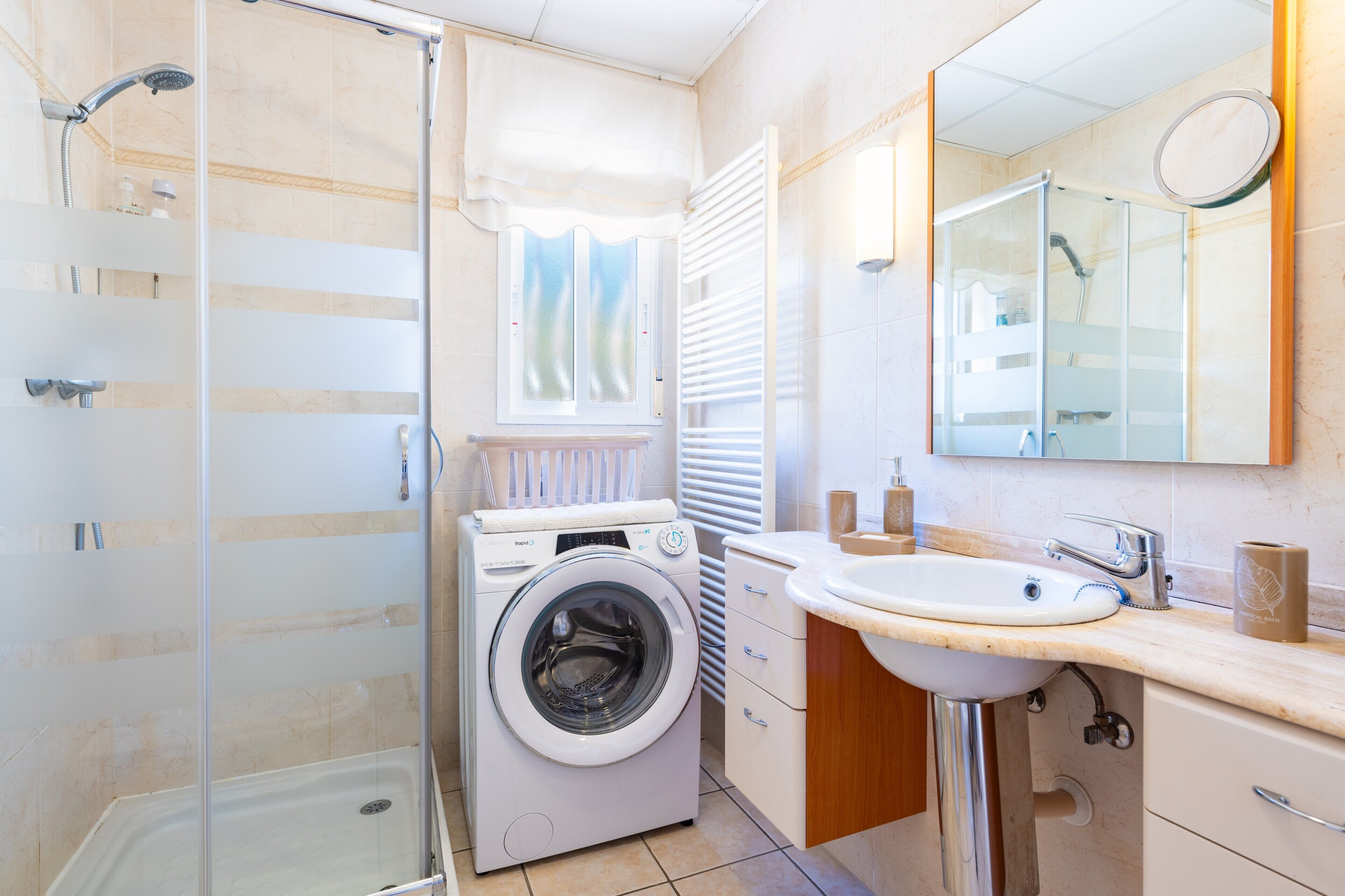 A compact bathroom with a washing machine, a sink, a mirror, and a shower enclosure.
