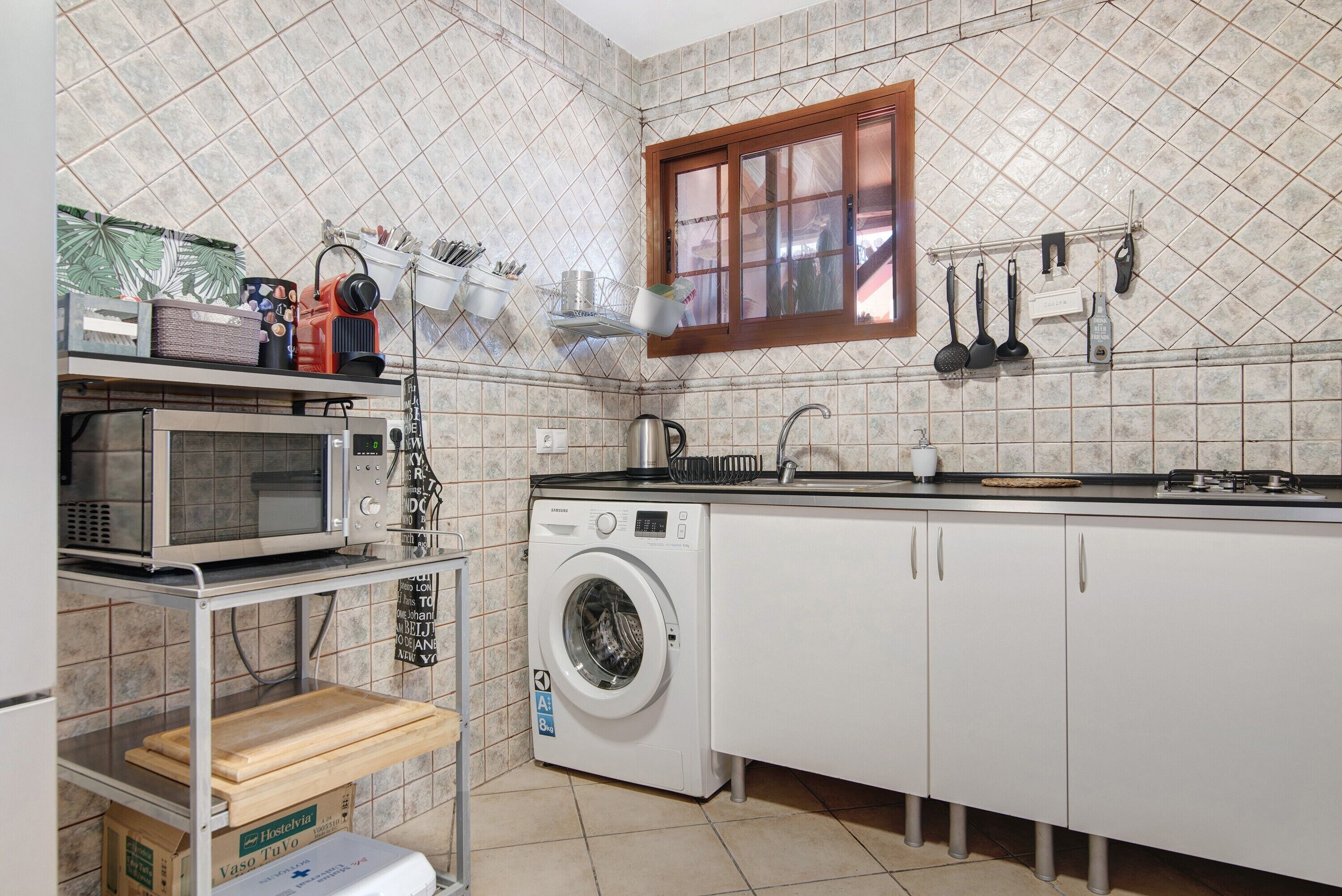 Cocina compacta con microondas, lavadora y ventana con utensilios colgando.