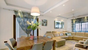 A modern living room with a wooden dining table, grey chairs, a beige sofa, and a large window with curtains. - Villa Dolce Vita Tenerife Sur with Sea Views, Private Pool, Terraces, and Wi-Fi (Adeje)
