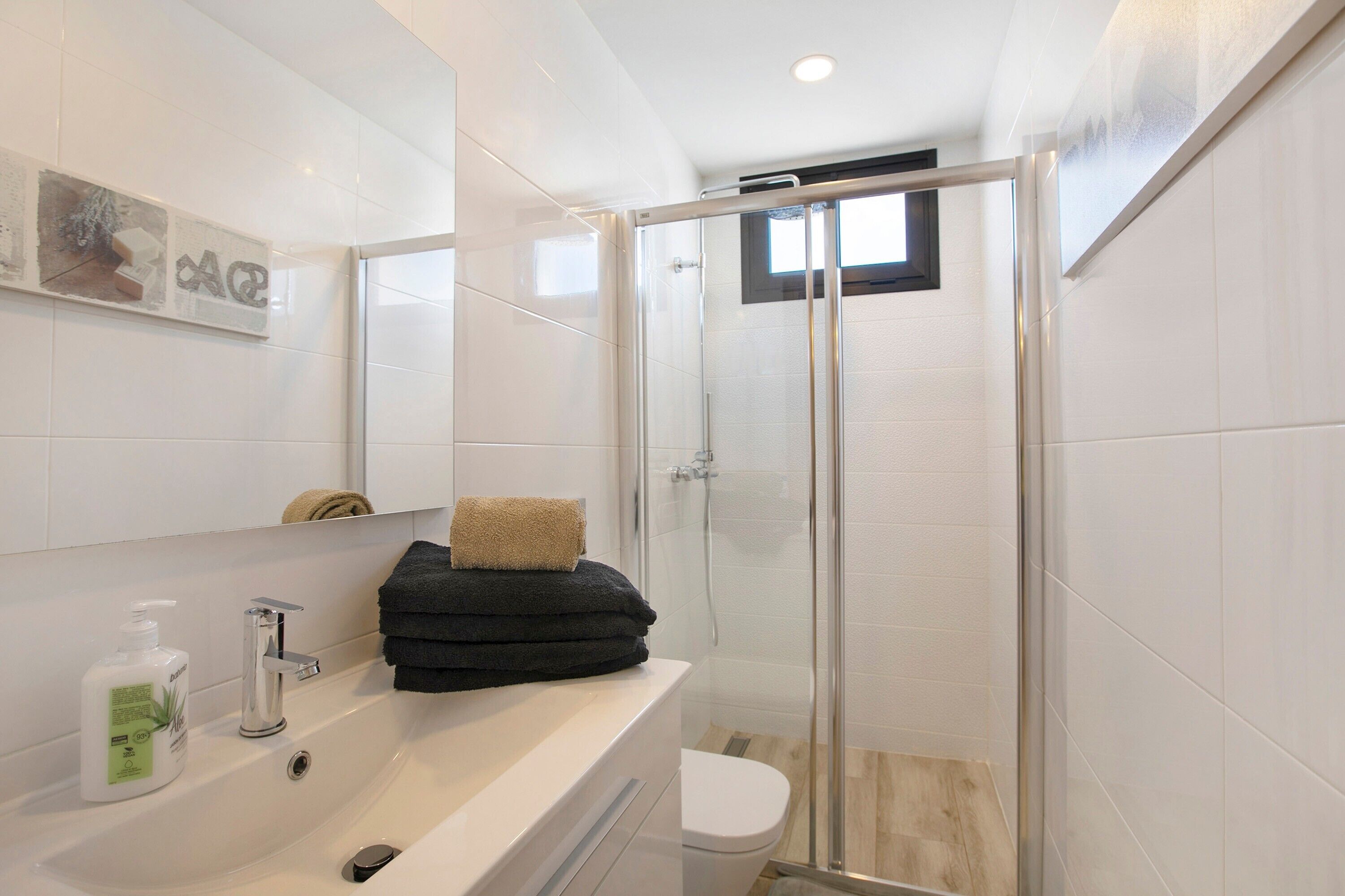 A bathroom with a glass shower enclosure, a white sink with a bottle of soap, and a towel stacked on the counter.