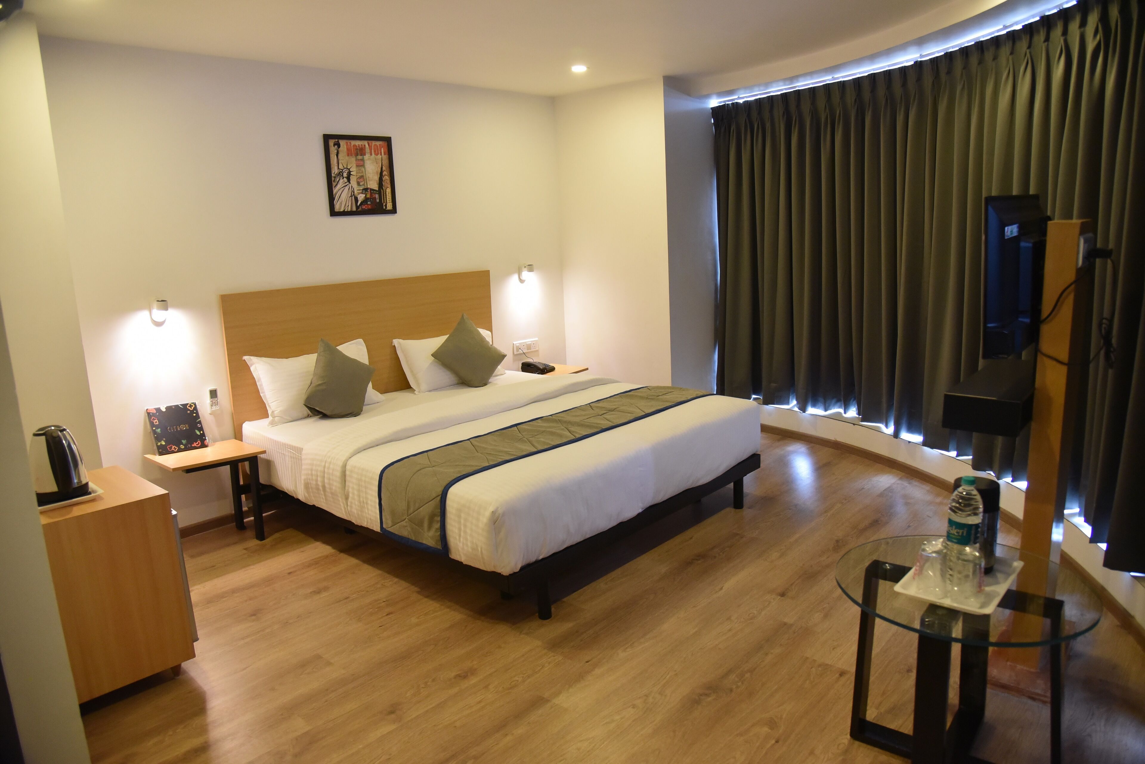 Quarto de hotel com cama, mesas de cabeceira, uma mesinha com uma garrafa, televisão e cortina.
