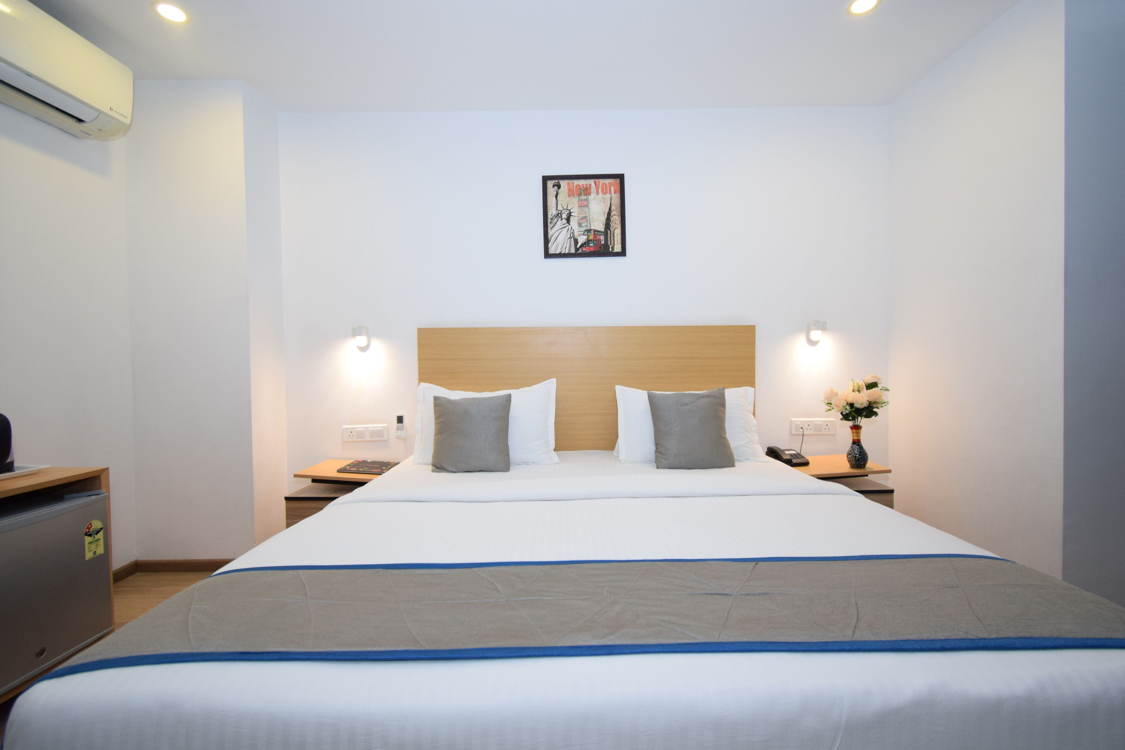 A hotel room with a large bed, a wooden headboard, two bedside tables with lamps, a wall-mounted air conditioner, and a framed picture above the bed.