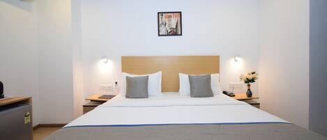 A hotel room with a large bed, a wooden headboard, two bedside tables with lamps, a wall-mounted air conditioner, and a framed picture above the bed.