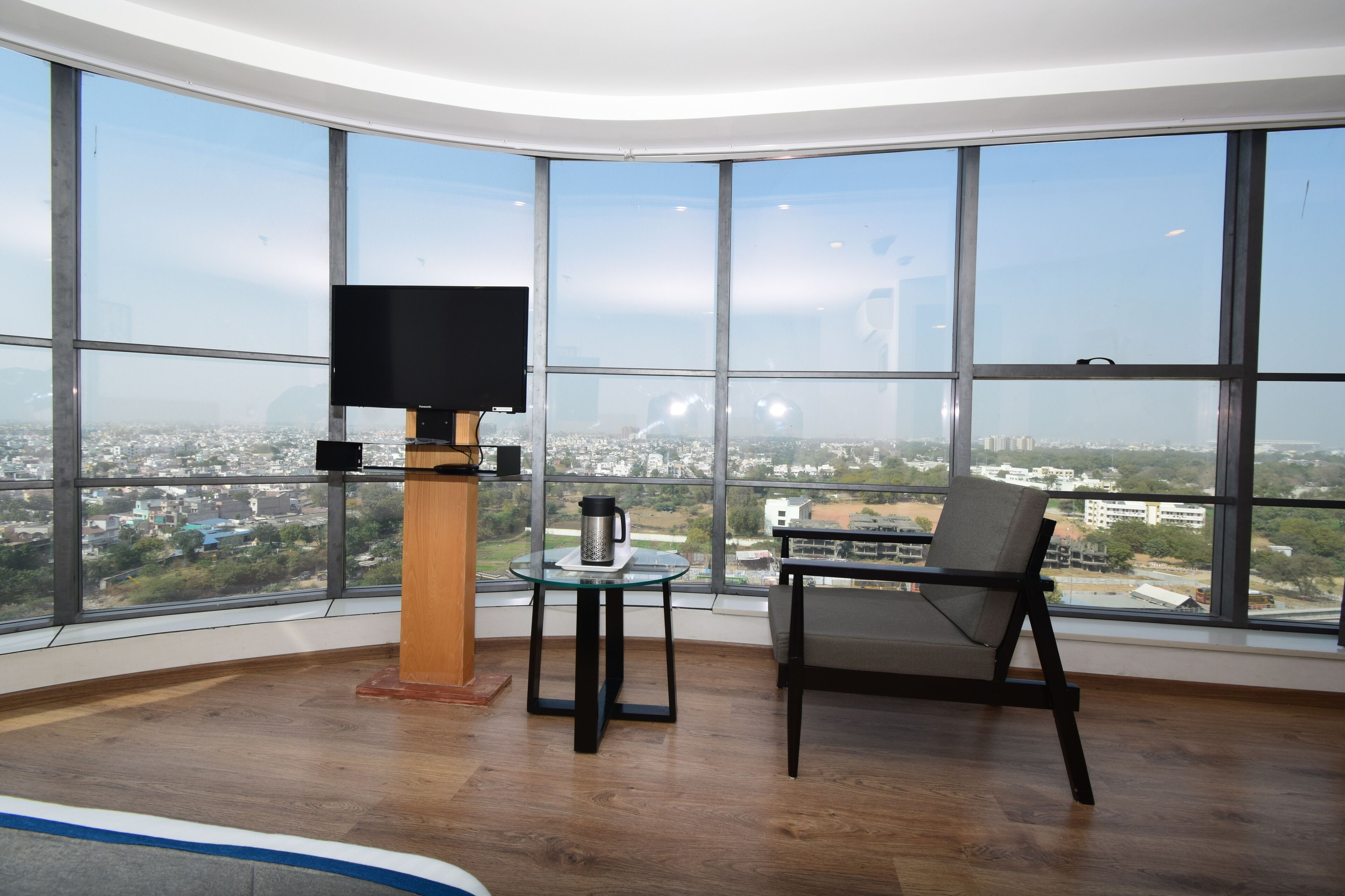 A spacious room with a large window offering a city view, a flat-screen TV, a small table with a coffee pot, and a comfortable chair.