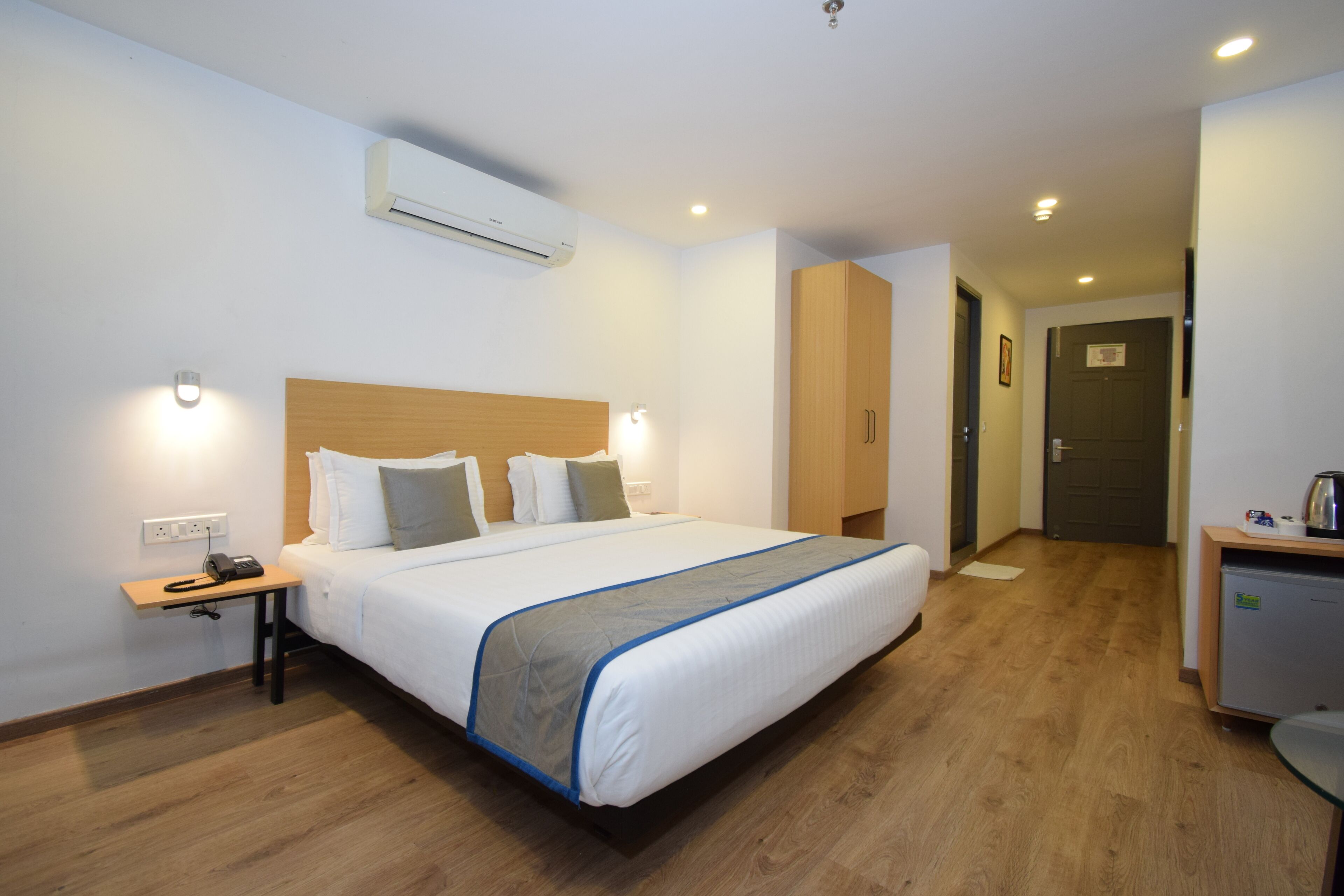 A hotel room with a large bed, bedside table with a phone, an air conditioning unit, a wooden wardrobe, and a kitchenette area.