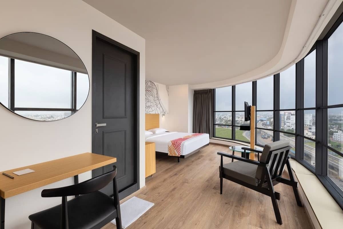 A modern hotel room with a bed, a desk, a chair, and large windows offering a city view.