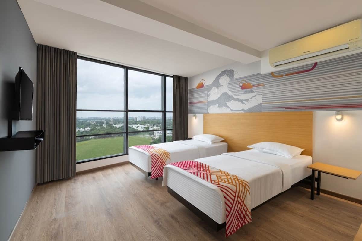 A hotel room with two beds, a large window with a city view, a mounted TV, and an air conditioning unit.