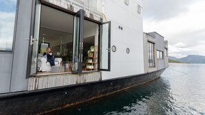 A modern houseboat with an open deck, a seating area with a sofa, and a plant shelf.